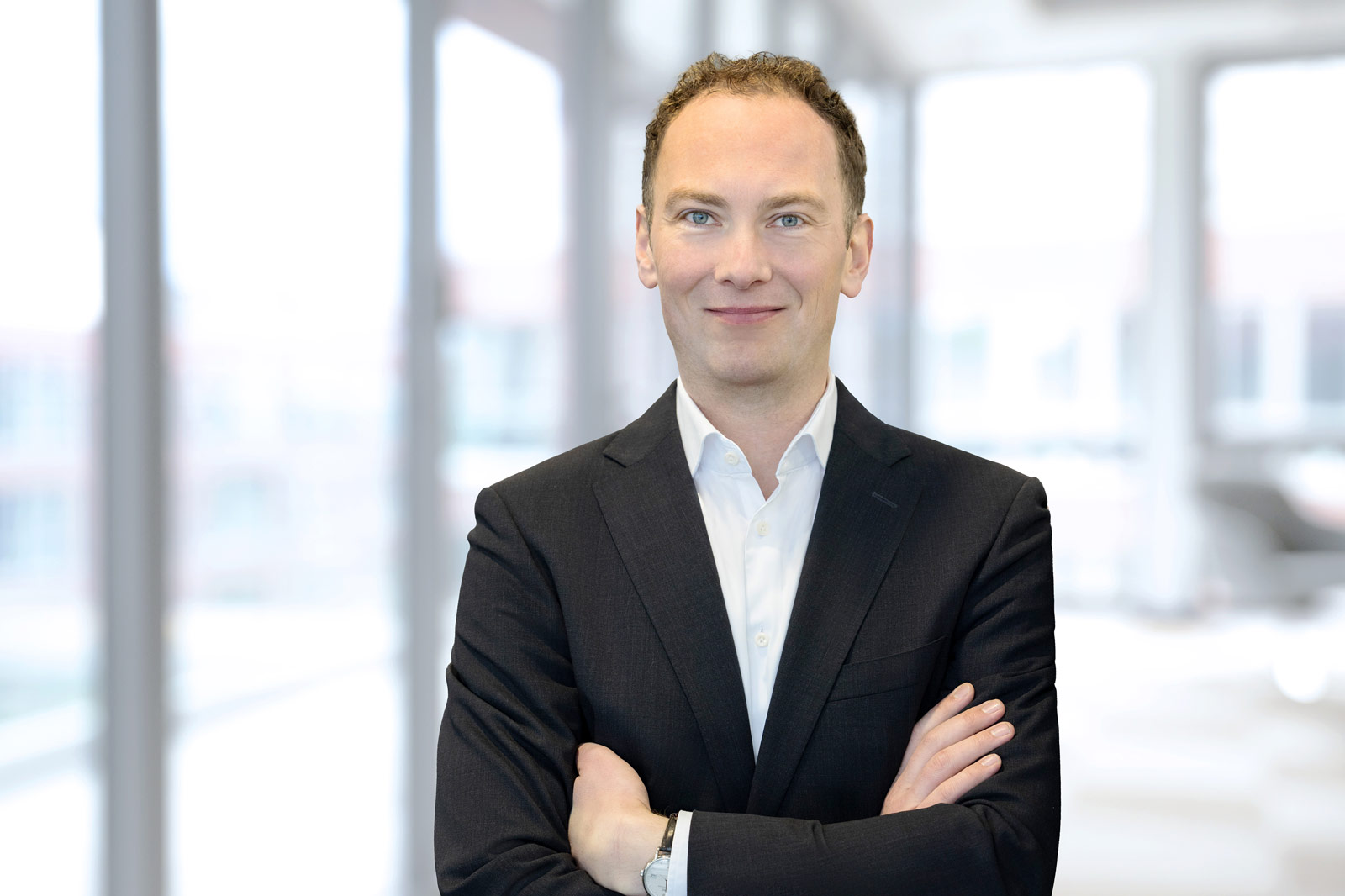 Ein Mann in einem dunklen Anzug steht mit verschränkten Armen in einem modernen, hellen Büro. Portrait von Tobias Keitel.
