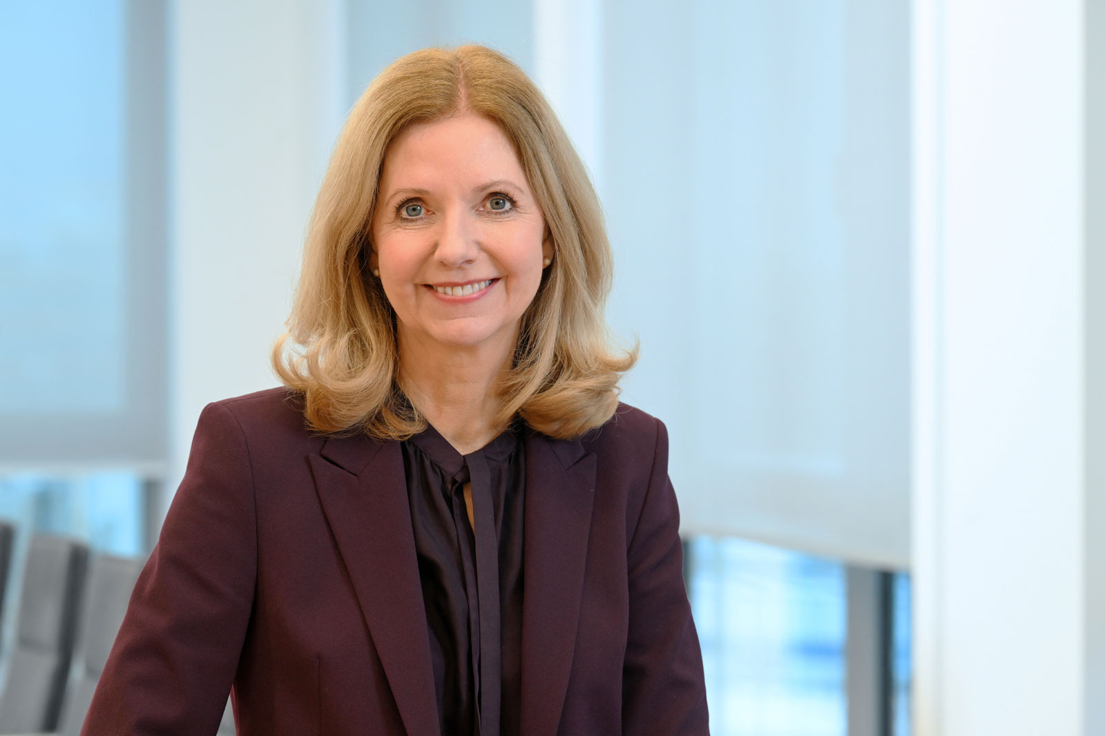 A woman with long, wavy blonde hair wears a dark blazer and stands in a modern office setting. Portrait of Katja van Doren.