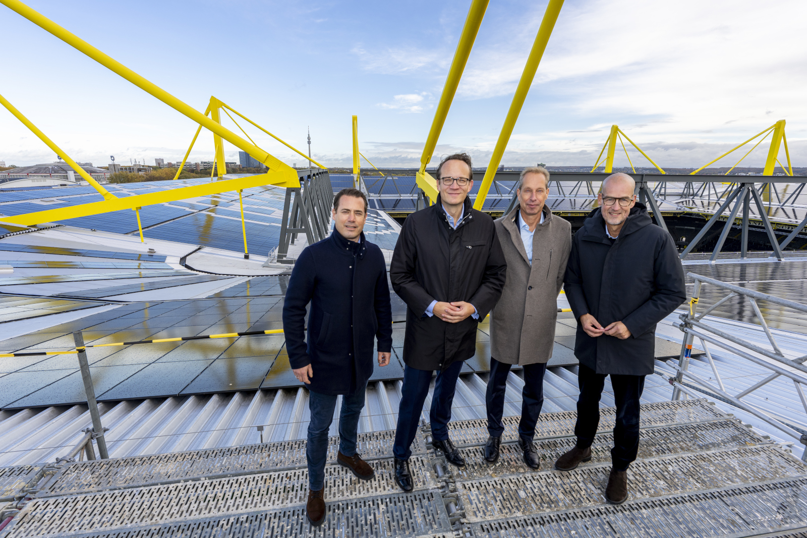 Four individuals stand on a rooftop with solar panels and yellow supports under a clear sky.