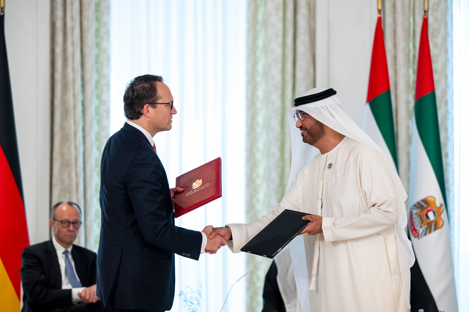 Two men shake hands during a signing ceremony with the flags of Germany and the UAE in the background.