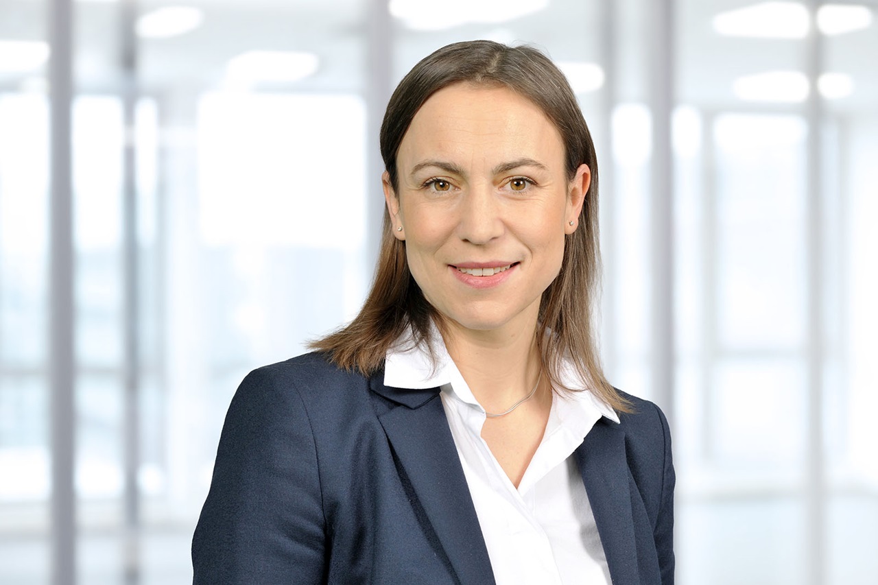 Eine beruflich gekleidete Frau mit schulterlangen braunen Haaren, dunkelblauem Blazer, weißer Hemdbluse und dünner silberner Kette in hellem Büro. Portrait von Sandra Dettmer.