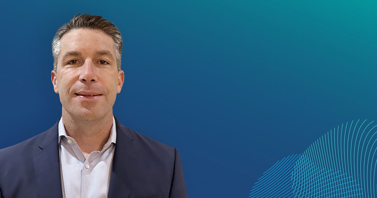 A man in a suit stands against a gradient blue background with abstract wave patterns.