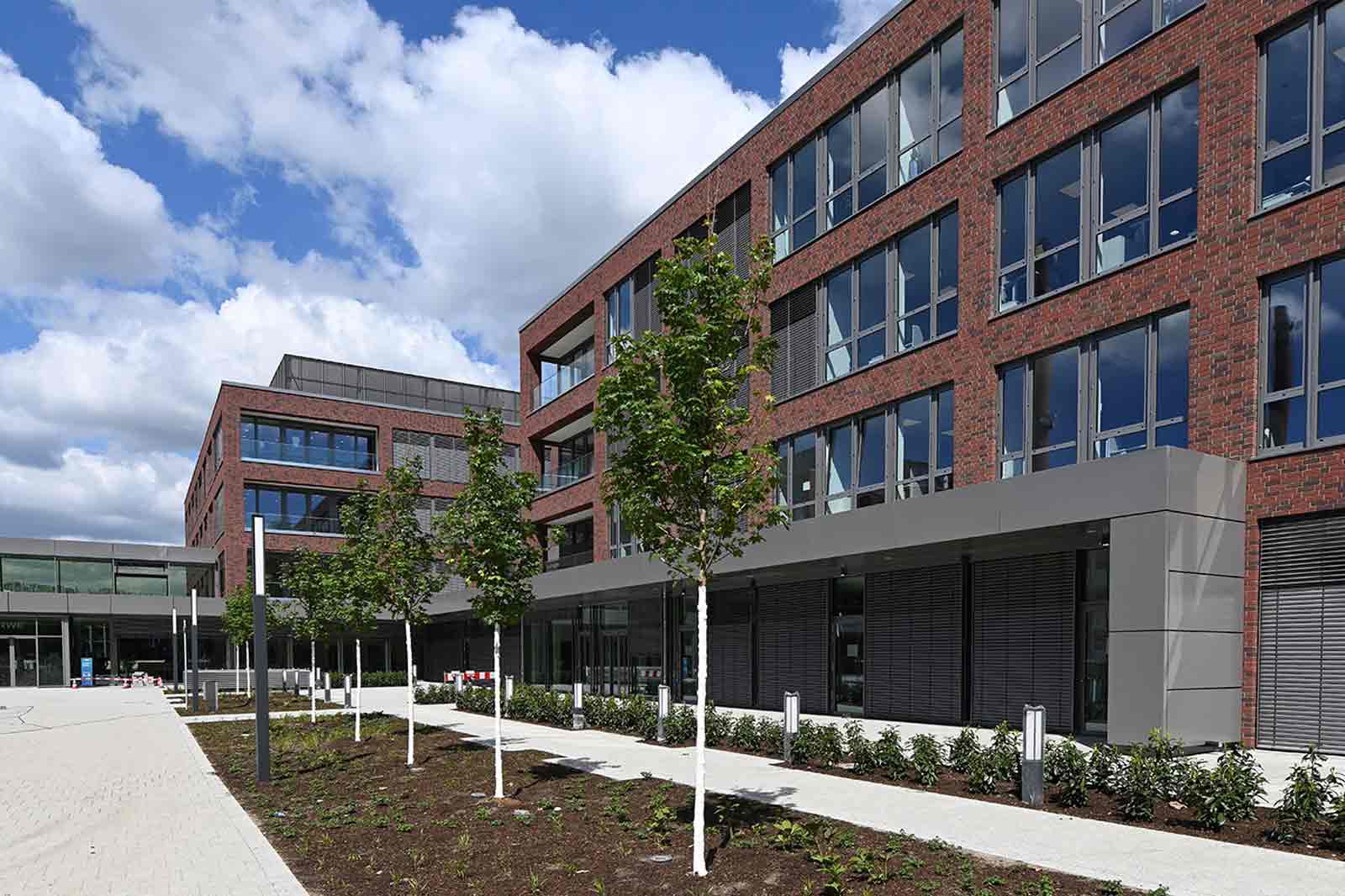 Ein modernes Gebäude mit roten Ziegeln, großen Fenstern und Bäumen im Vorgarten unter einem blauen Himmel.