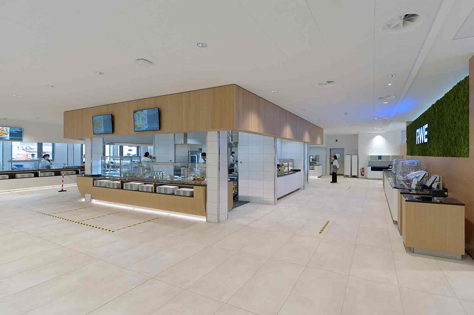 Moderne Cafeteria mit eleganten Holzelementen, verschiedenen Essensstationen und digitalen Menüs.