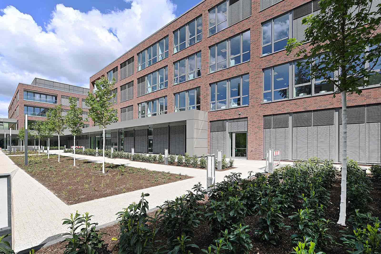 Moderne Gebäude mit großen Fenstern und begrünten Flächen. Bäume und Pflastersteige umgeben den Eingangsbereich.