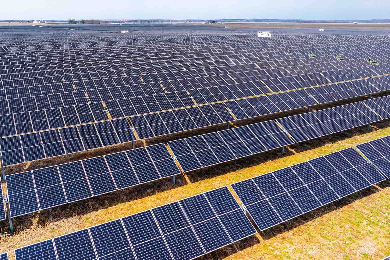Reihen dunkelblauer Photovoltaikmodule auf Metallgestellen bedecken flaches gelblich-braunes Grasland und erstrecken sich in ordentlichen Gittern bis zum blassen Horizont.
