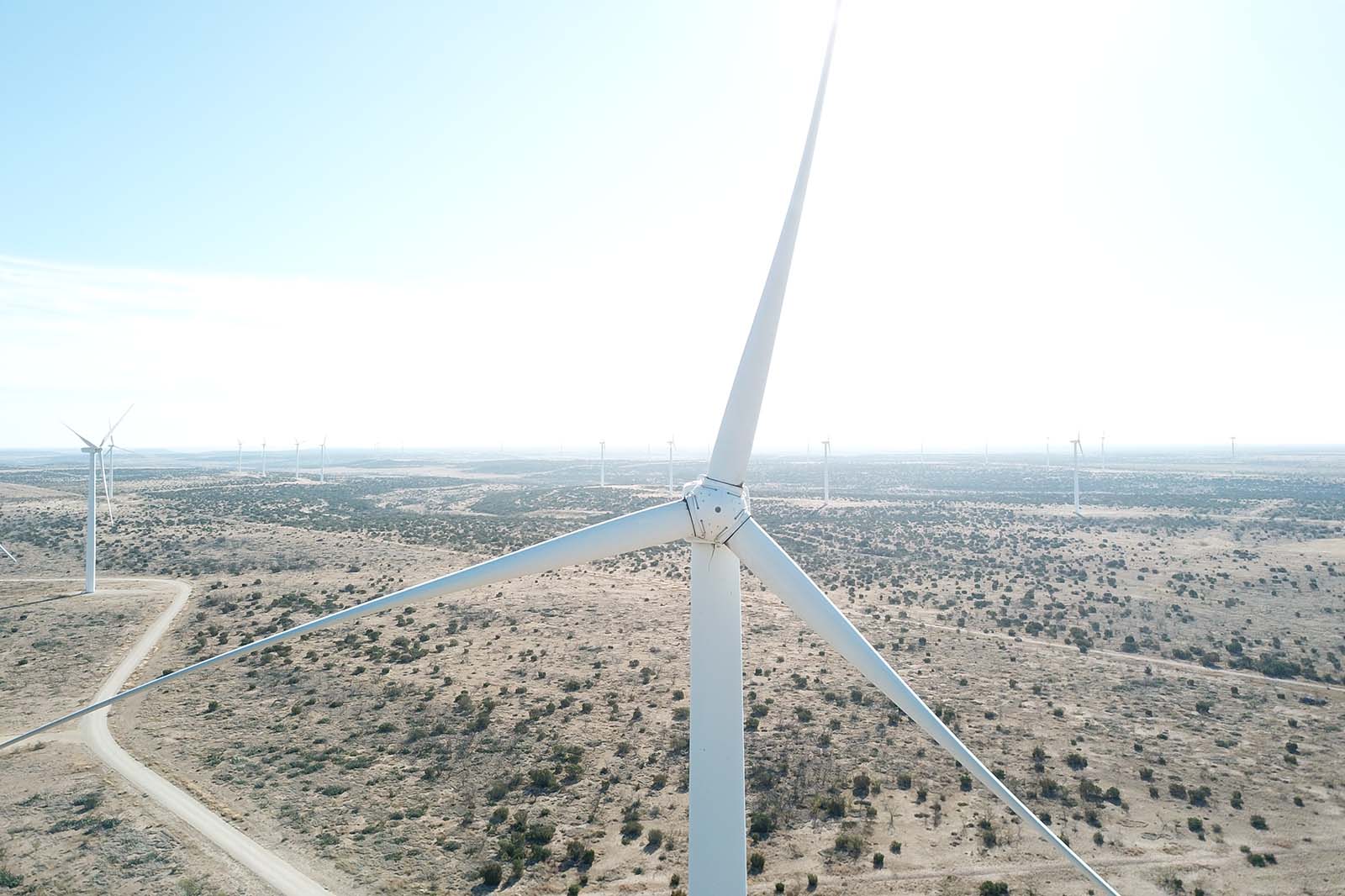 A close aerial view of a white wind turbine rotor and blades over a beige, scrubby desert with dirt roads and distant turbines.