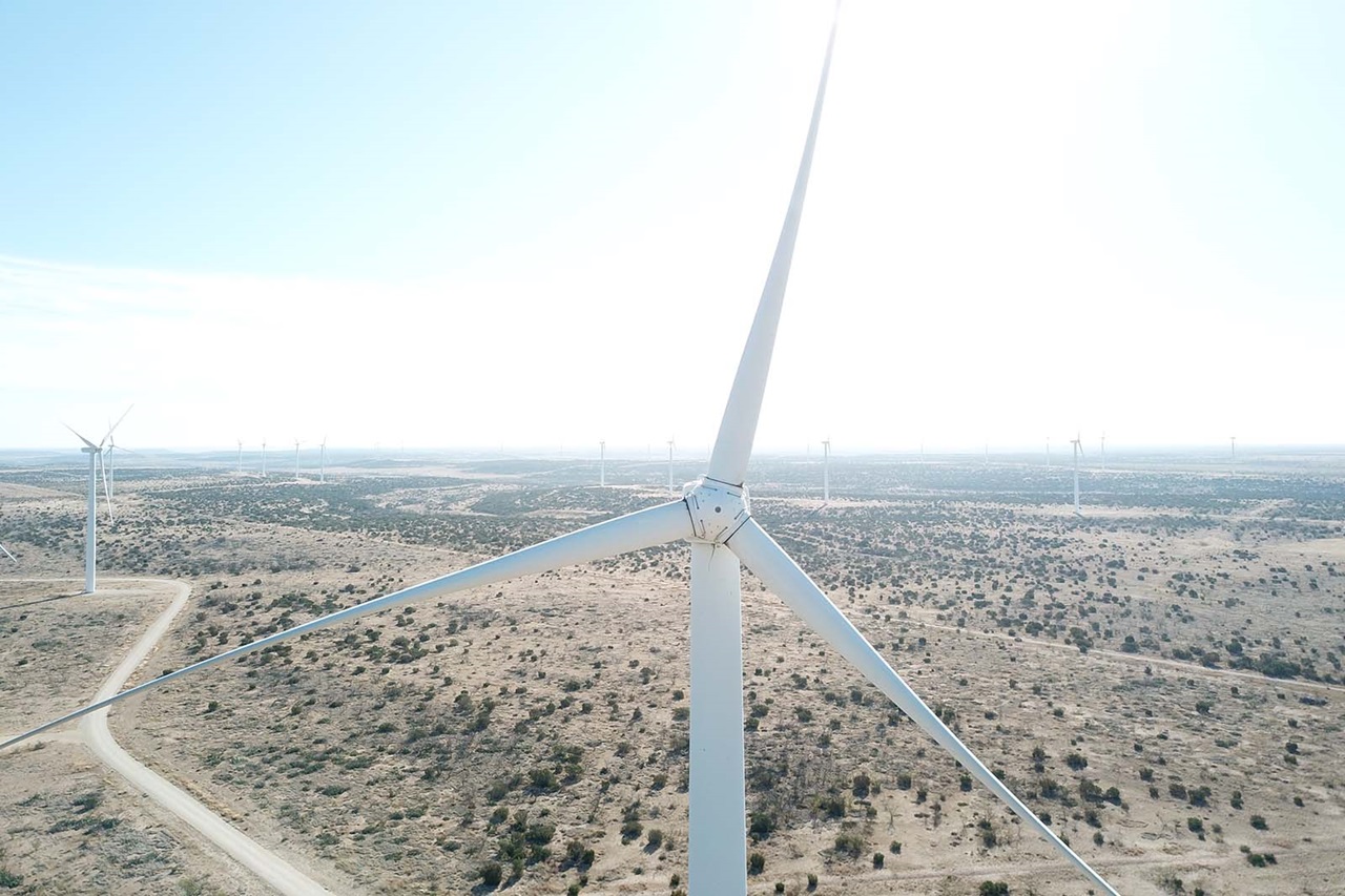 A close aerial view of a white wind turbine rotor and blades over a beige, scrubby desert with dirt roads and distant turbines.
