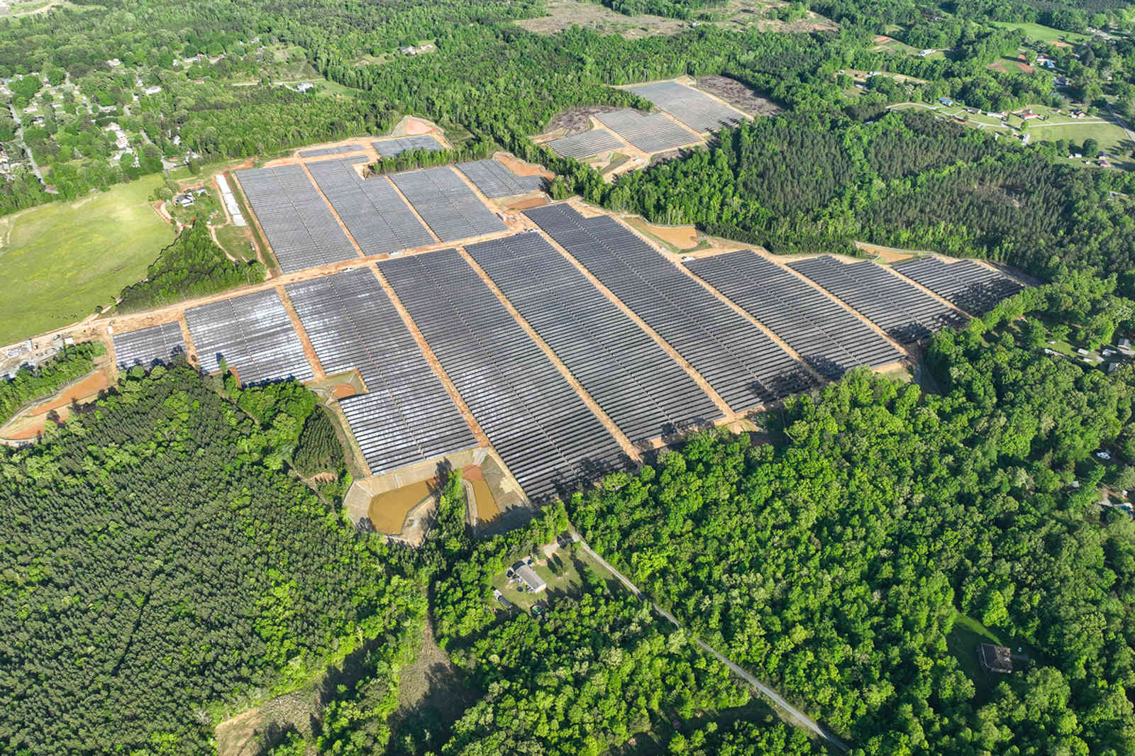 Aerial view of a large solar farm surrounded by greenery and residential areas, featuring numerous solar panels on rows.