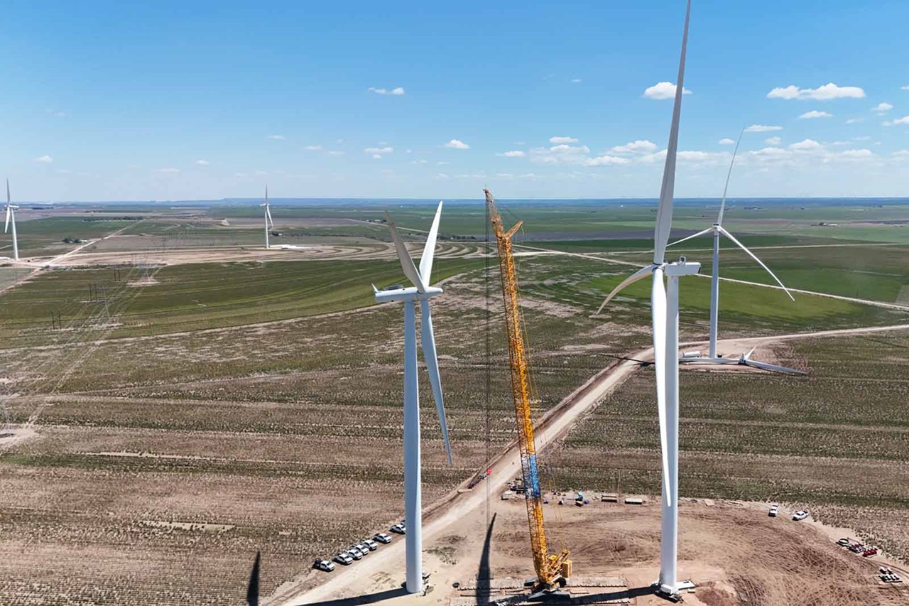 Aerial view of wind turbines in a green landscape, with a tall crane assembling one turbine and blue skies above.