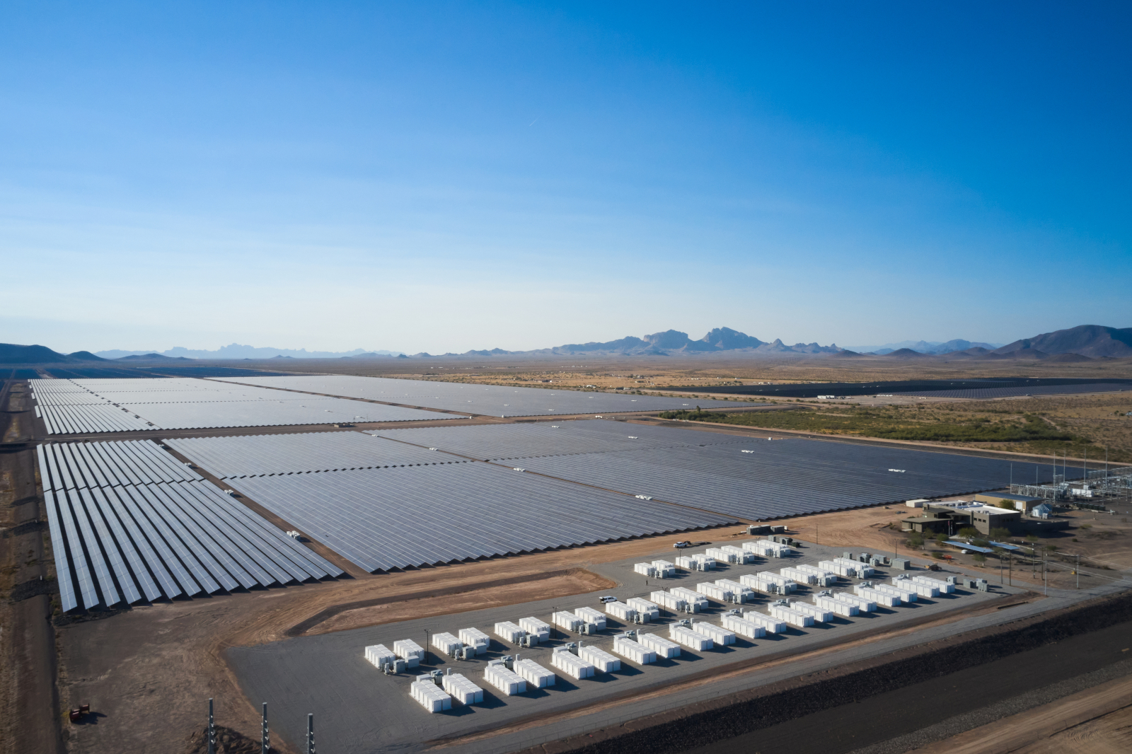A vast solar farm with rows of panels under a clear blue sky and mountains in the background. Storage units are visible.