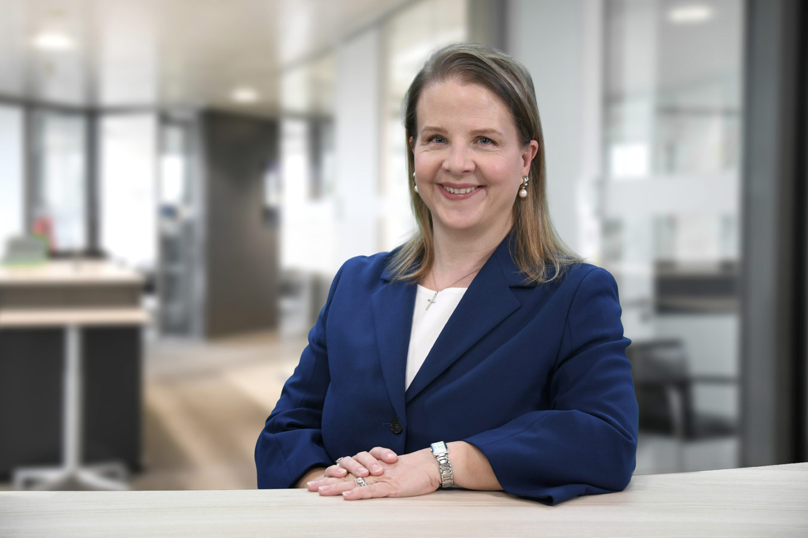 Sabine Augustin in einem blauen Blazer sitzt an einem Tisch in einem modernen Büro. Der Hintergrund ist unscharf.
