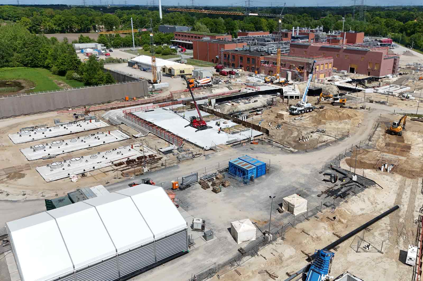 Eine Baustelle mit verschiedenen Maschinen, Betonplatten und einem großen Zelt im Vordergrund. Es gibt Bäume im Hintergrund.