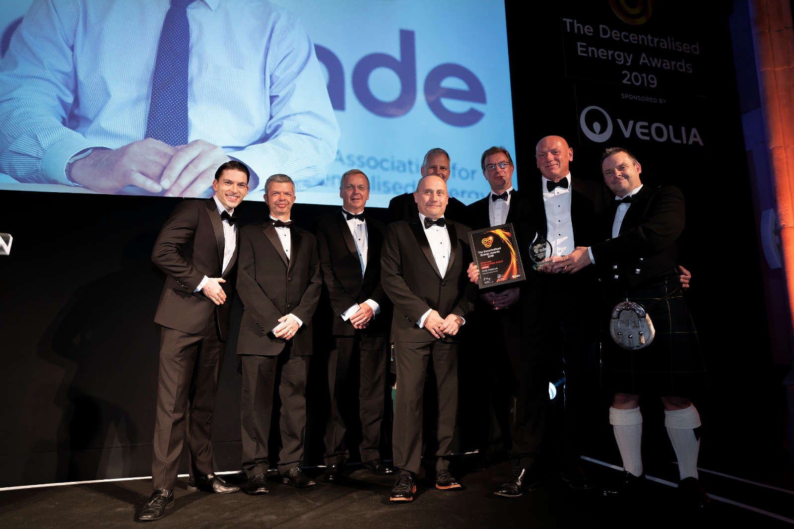 A group of men in formal attire, including one in a kilt, holding an award on stage at the Decentralised Energy Awards 2019.