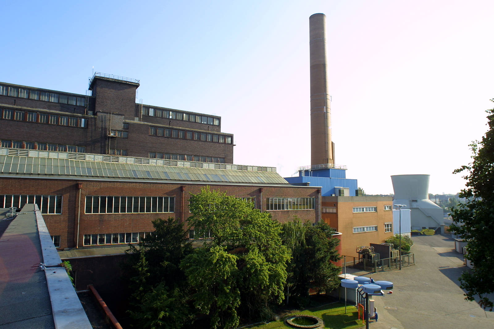 Ein großes Industriegebäude mit einem hohen Schornstein und Kühlturm, umgeben von Grünanlagen und blauem Himmel.