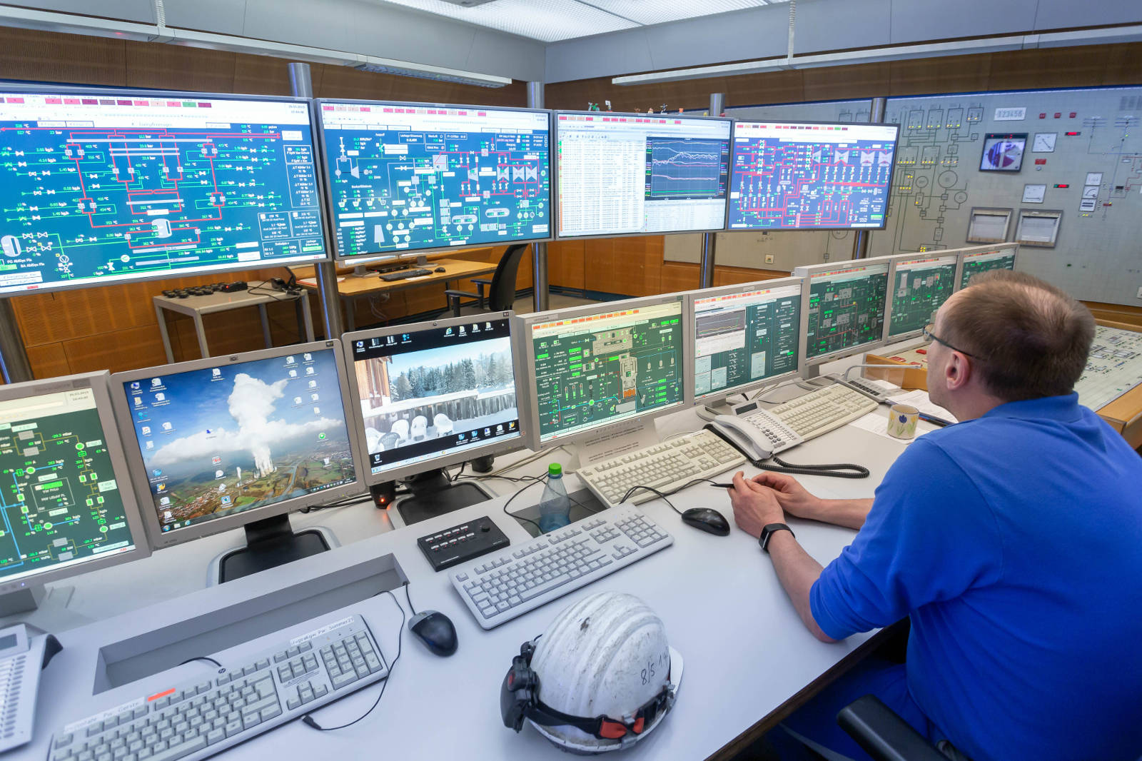 Ein Kontrollraum mit einer Person, die mehrere Bildschirme überwacht, die technische Daten und Grafiken anzeigen, mit einem Schutzhelm auf dem Tisch.