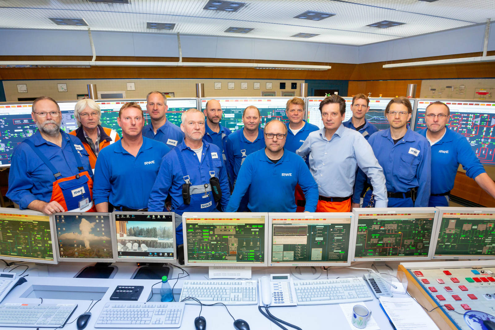 Eine Gruppe von Fachleuten in blauen Uniformen posiert in einem Kontrollraum mit Bildschirmen und Geräten.