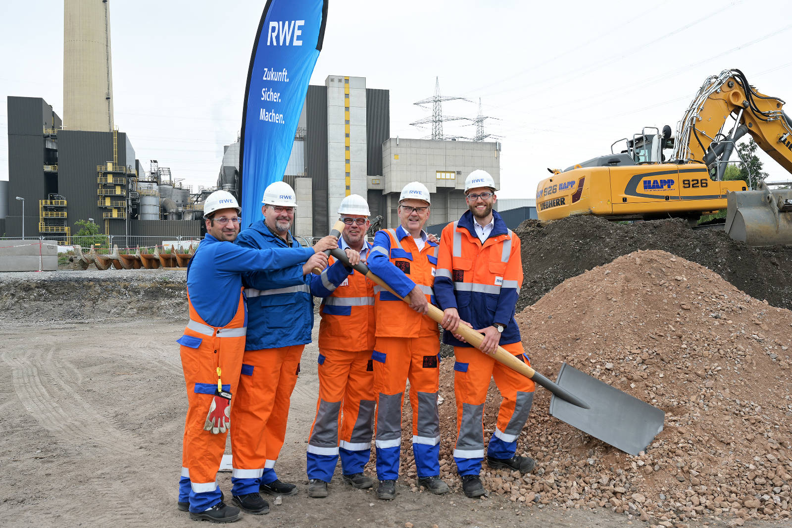 Fünf Bauarbeiter stehen gemeinsam, tragen Sicherheitskleidung und stehen auf einer Baustelle mit Maschinen und einem großen Kieshaufen.