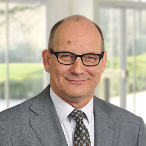 A man in a grey suit and patterned tie stands in a bright, modern setting with large windows.