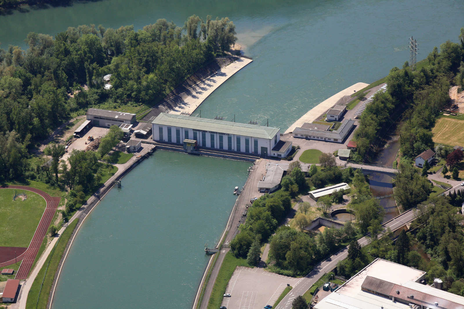 Luftaufnahme eines Damms und eines Kraftwerks an einem Fluss, umgeben von grünen Bäumen und Gebäuden.