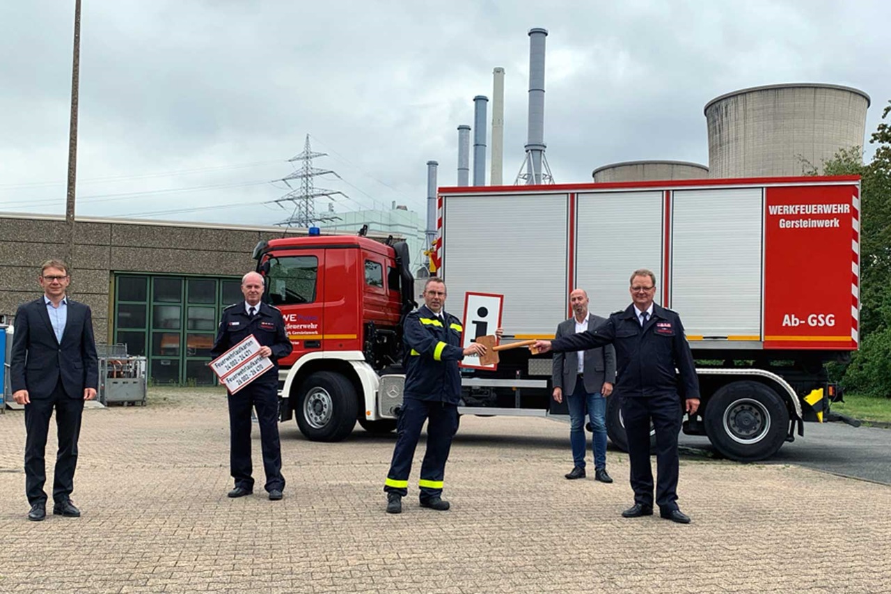 Eine Gruppe von Personen nimmt an einer Zeremonie in der Nähe eines Feuerwehrfahrzeugs an einem Industriestandort teil.