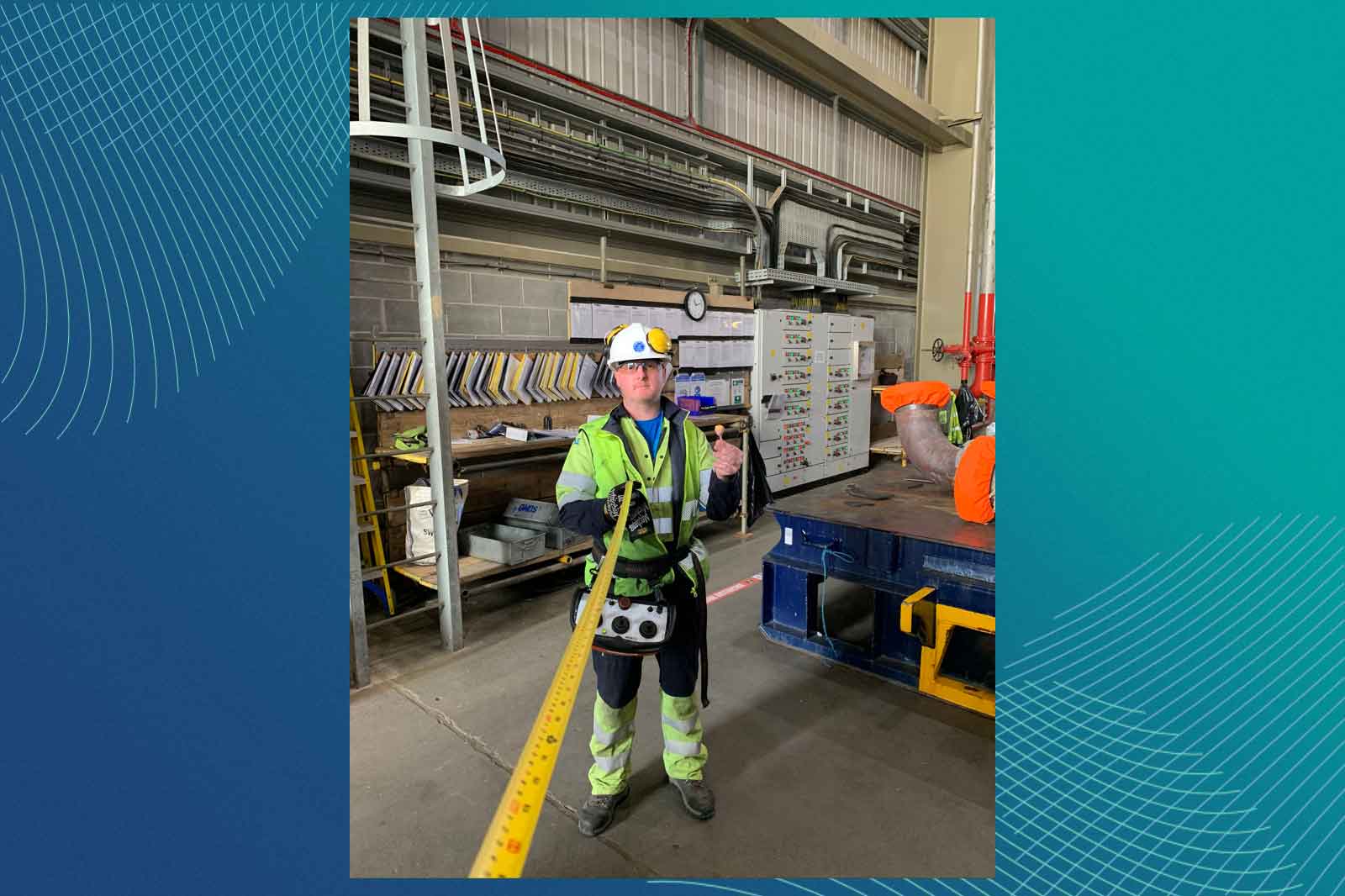 A worker in a safety vest stands in a warehouse, holding a measuring tape next to a workbench and equipment.