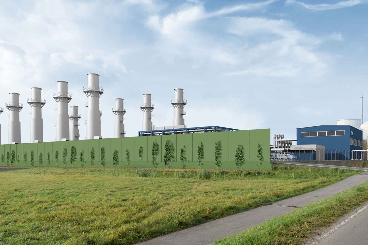 A modern industrial facility with tall chimneys, surrounded by a green privacy wall and grassy areas.