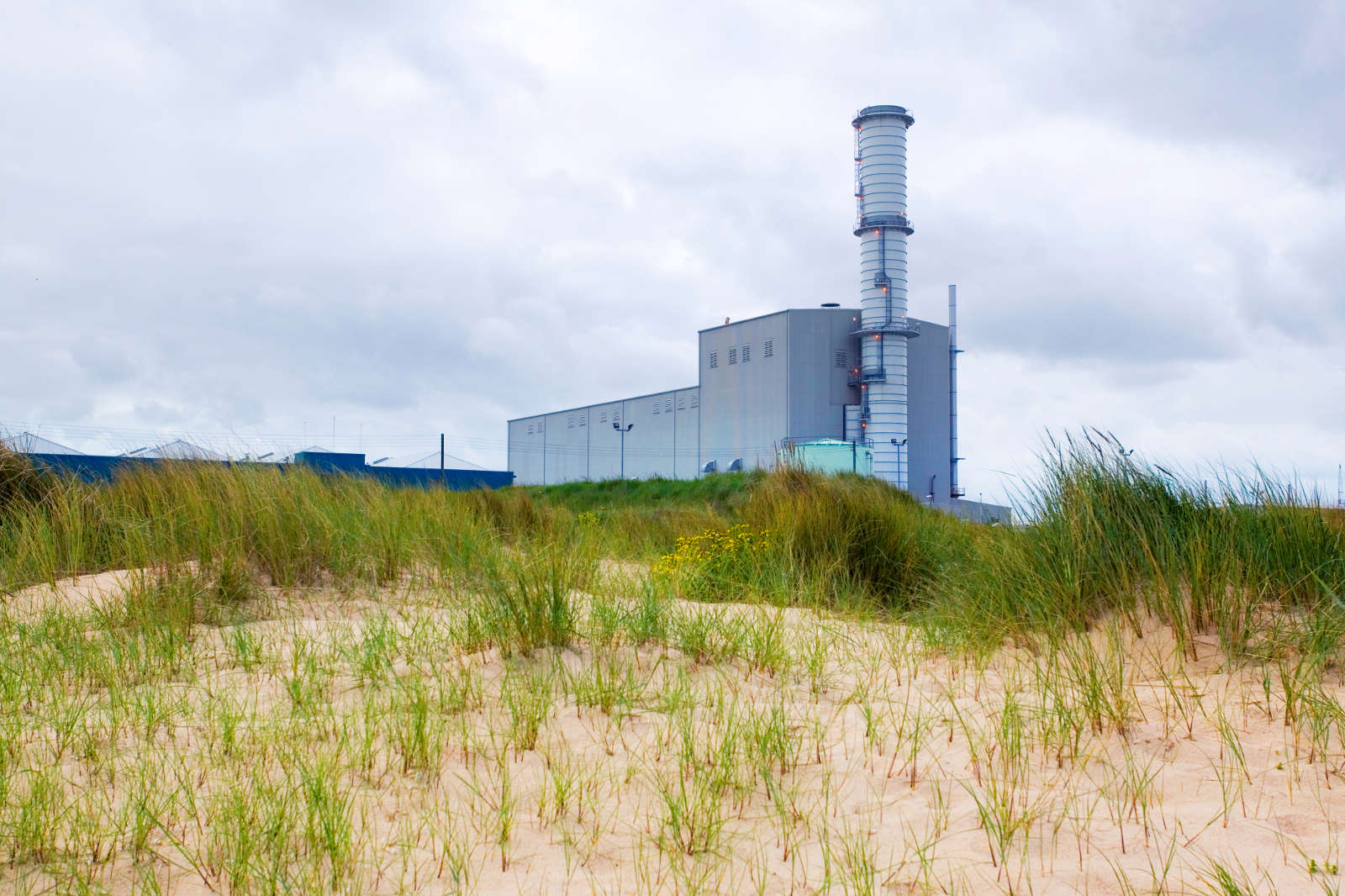 A power plant is visible behind sandy dunes and green grass under a cloudy sky.