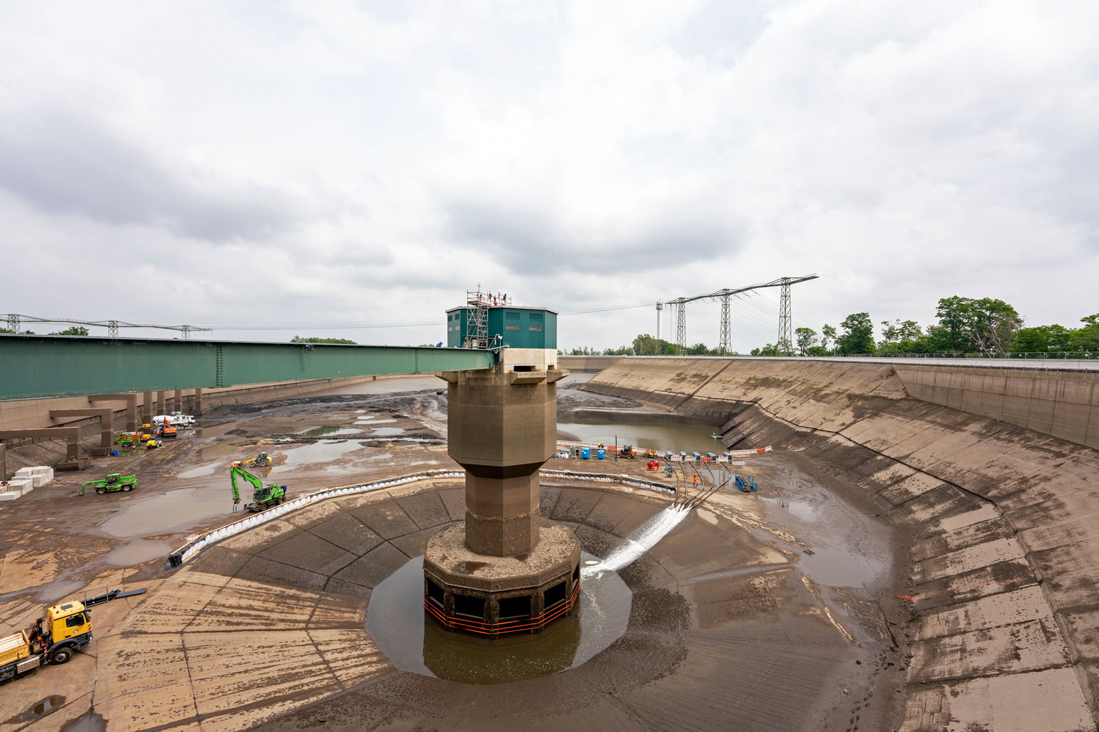 Ein Blick auf einen leeren Dammreservoir, der renoviert wird, mit schweren Maschinen und einem Bedienungsturm unter bewölktem Himmel.