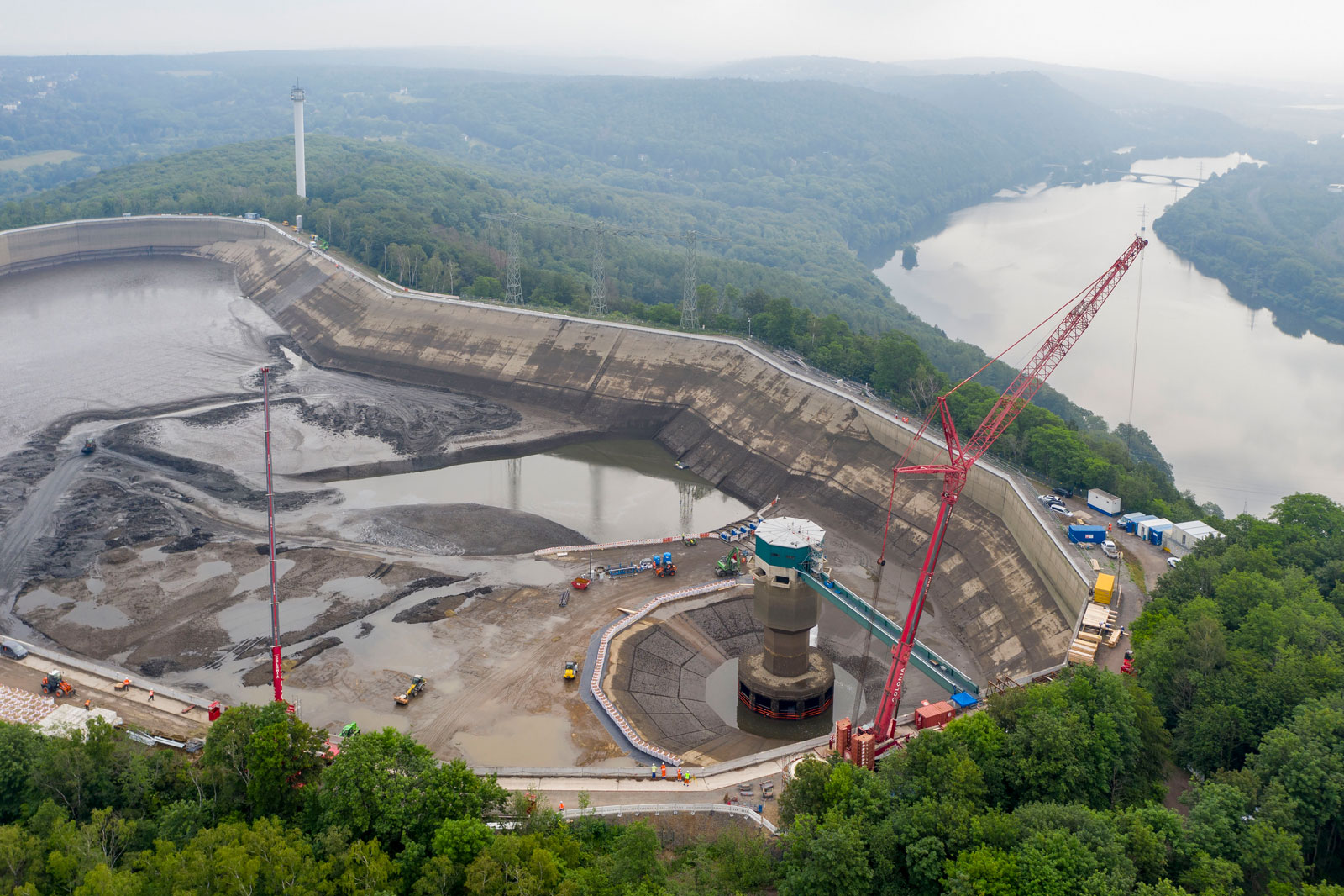 Luftaufnahme einer Baustelle an einem Damm mit Kränen, Maschinen und einem Fluss im Hintergrund.