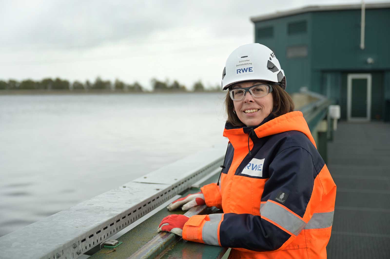 Eine Person in einer orangefarbenen Sicherheitsjacke und einem Helm steht am Wasser und beobachtet die Umgebung.