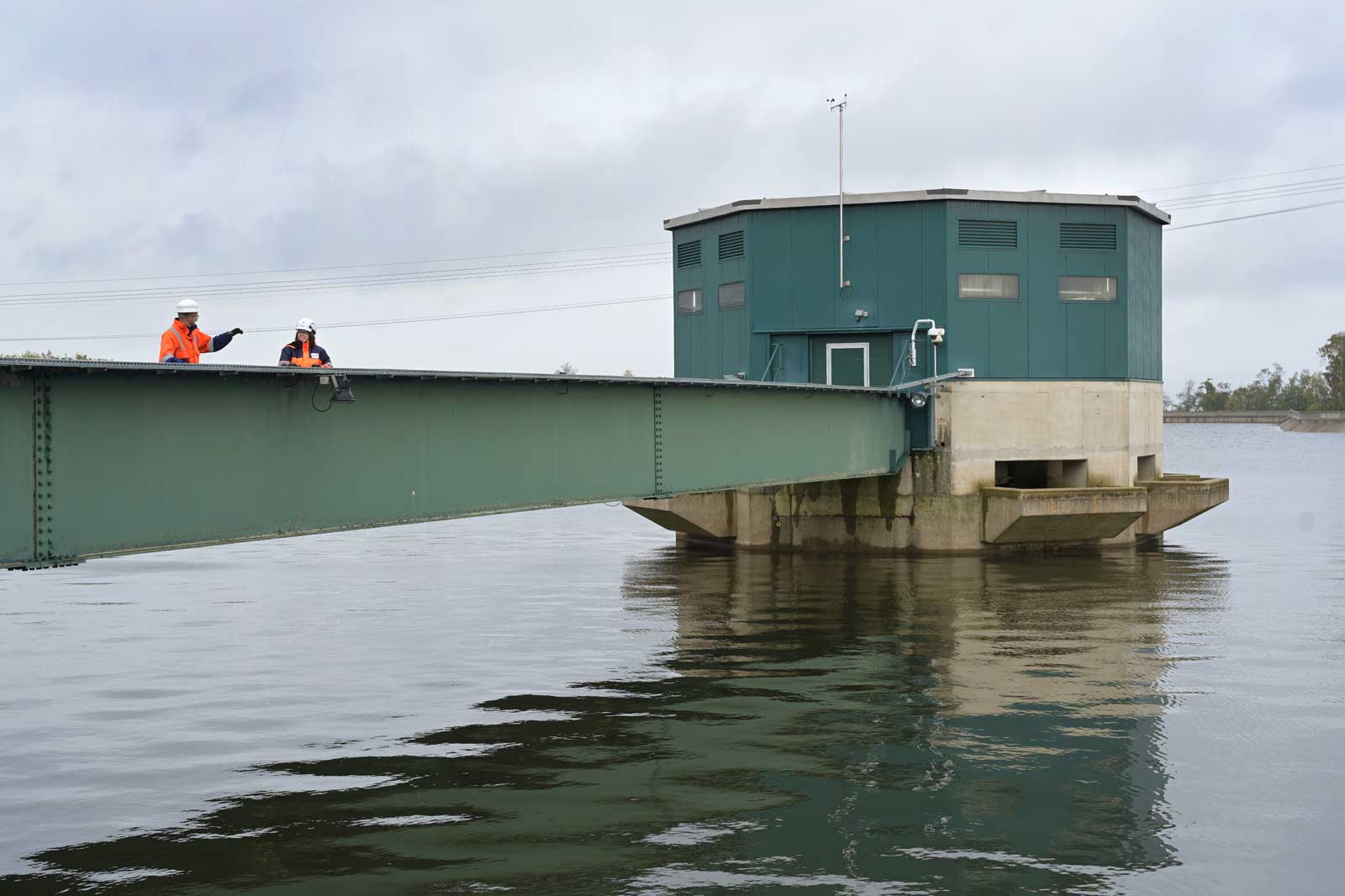 Ein grünes Kontrollgebäude auf Stelzen über Wasser, mit zwei Arbeitern in orangenen Jacken, die die Struktur inspizieren.
