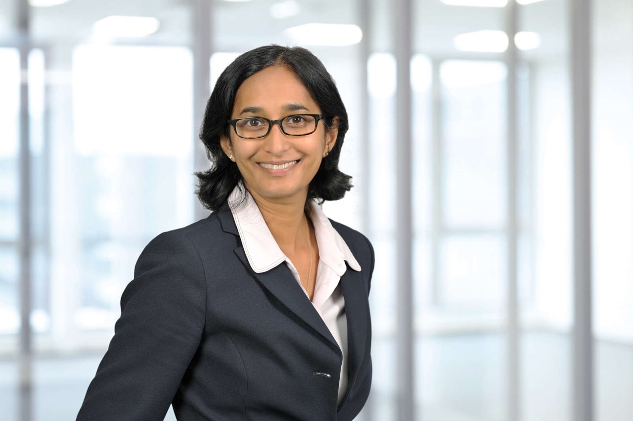 A woman in a formal suit stands in a modern office environment with large windows in the background.