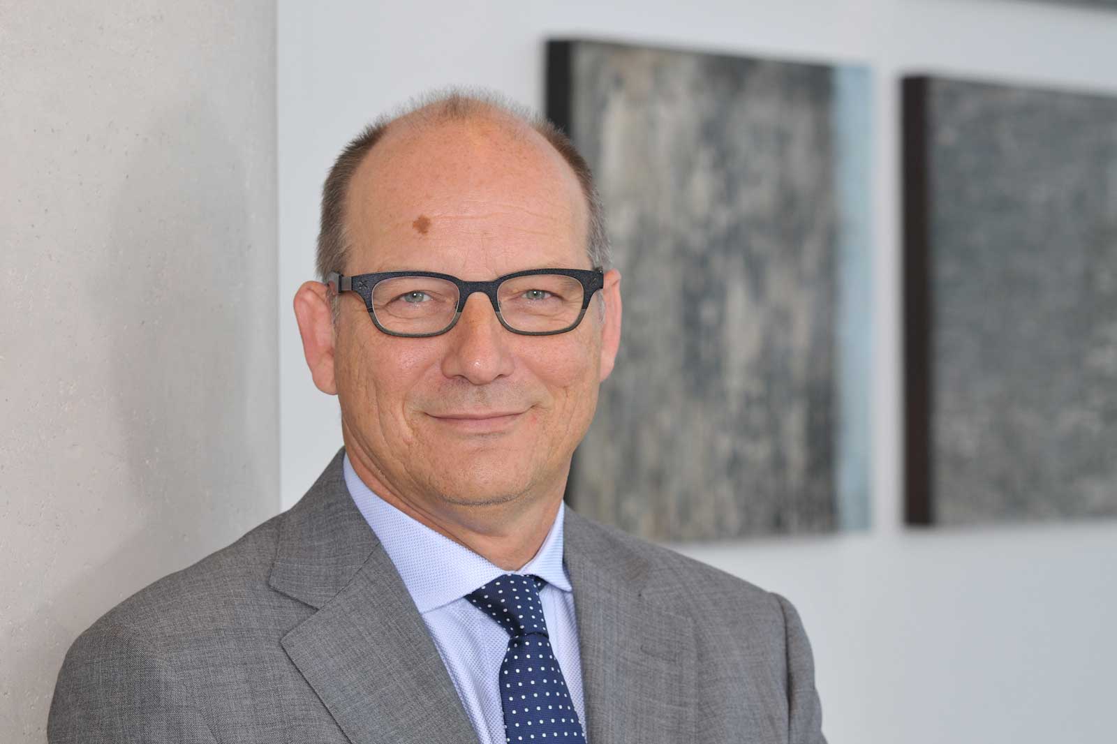 A man in a suit stands beside a grey wall with artworks in the background.