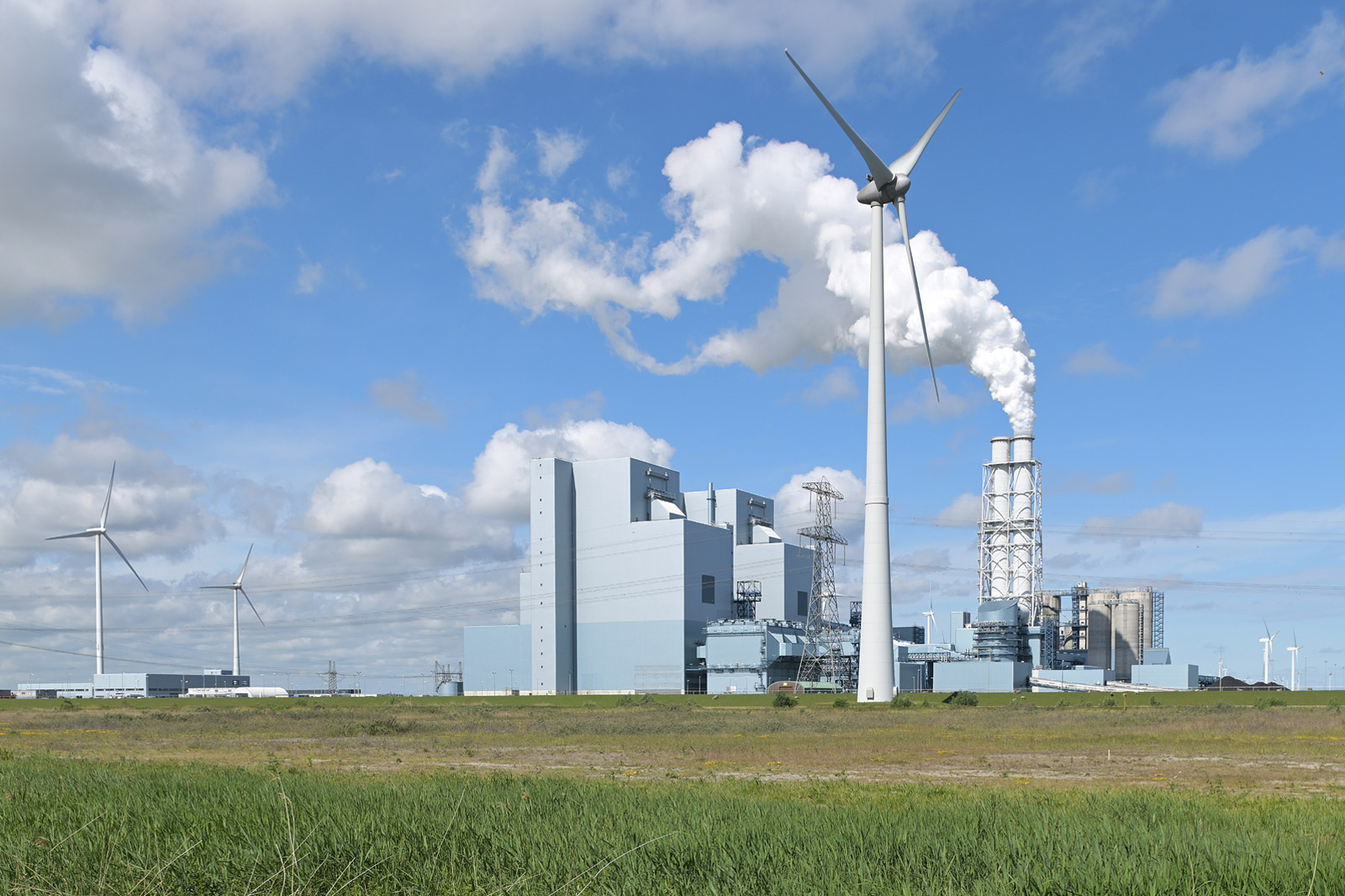 An industrial complex featuring wind turbines and smoking chimneys against a blue sky with clouds.
