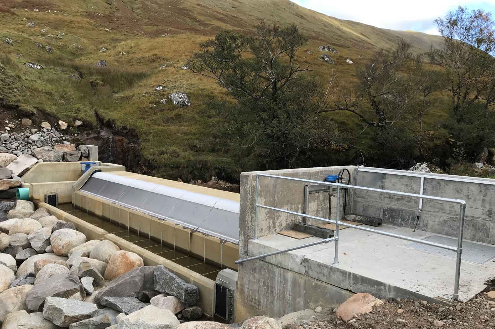 A modern water infrastructure with a concrete channel, surrounded by rocks and hill slopes, featuring a guard rail and plumbing equipment.