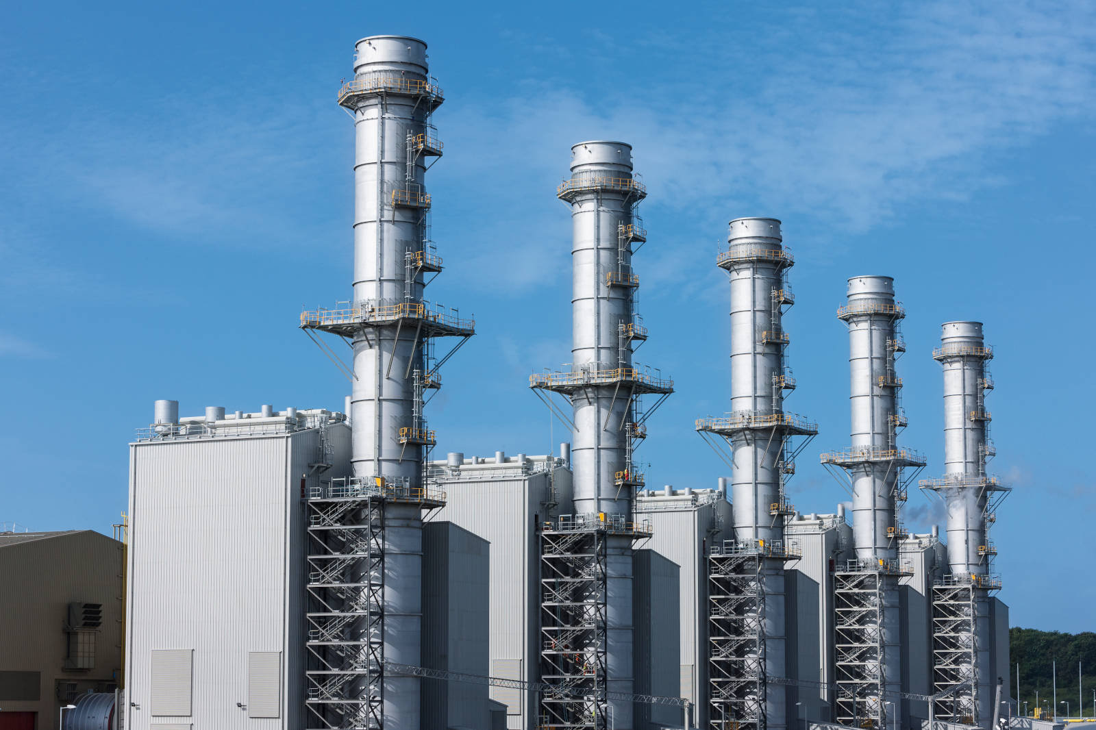 Five tall industrial chimneys stand against a blue sky. The buildings are made of silver metal and feature staircases.