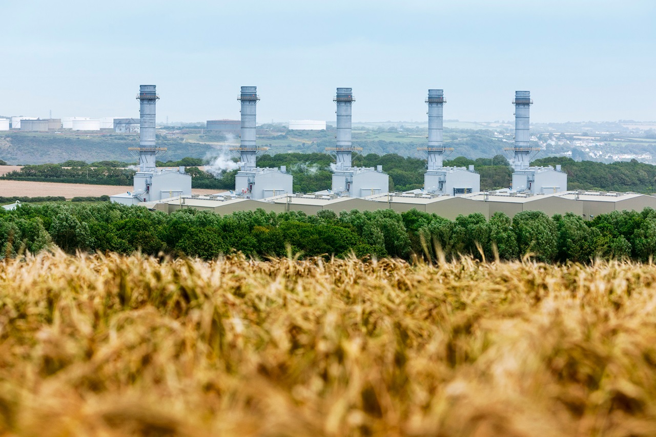 A landscape featuring industrial power plants with tall chimneys, surrounded by green trees and golden fields.