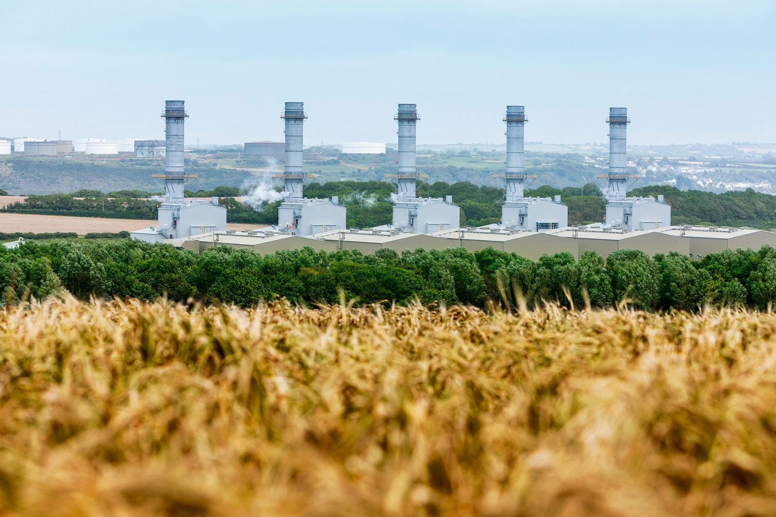 A landscape featuring industrial power plants with tall chimneys, surrounded by green trees and golden fields.