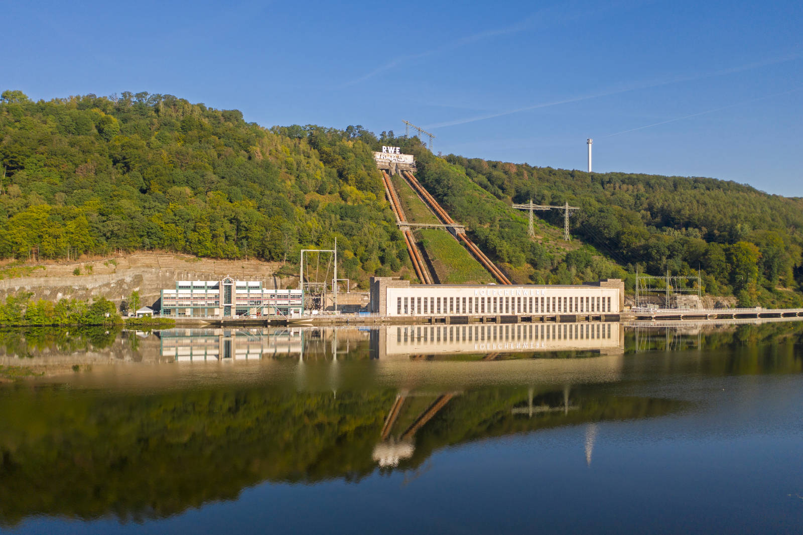 Ein Wasserkraftwerk neben einem ruhigen See, umgeben von grünen Hügeln und klarem blauen Himmel.