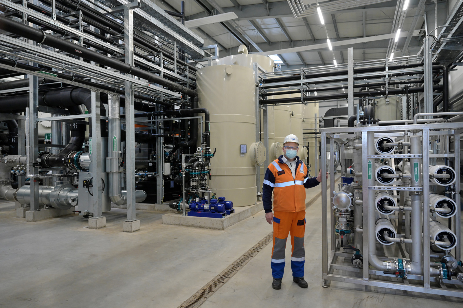 Ein Arbeiter in orange-blauen Overalls steht neben industriellen Anlagen in einer großen Einrichtung mit verschiedenen Rohren und Tanks.