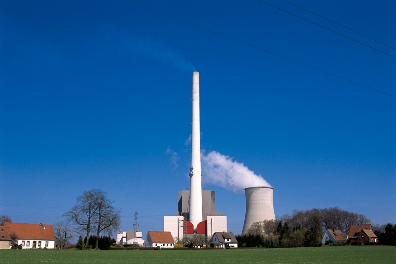 A factory with a tall chimney and a cooling tower is depicted against a clear blue sky. Several houses are visible in the foreground.