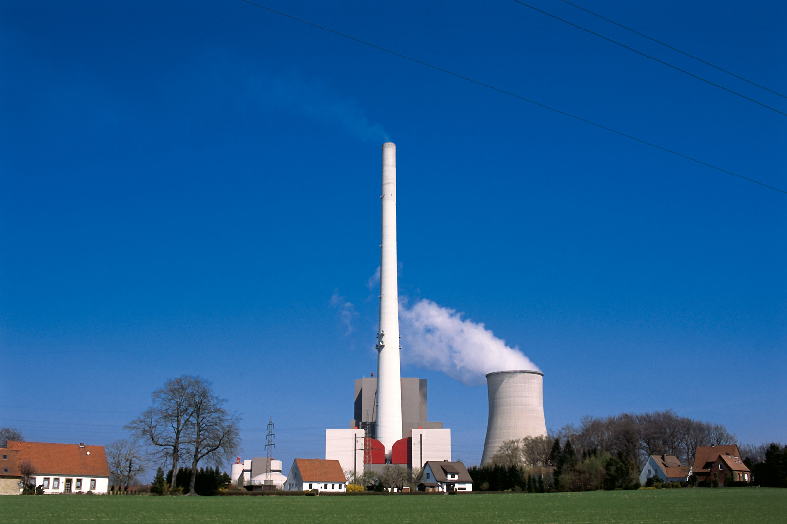 A factory with a tall chimney and a cooling tower is depicted against a clear blue sky. Several houses are visible in the foreground.