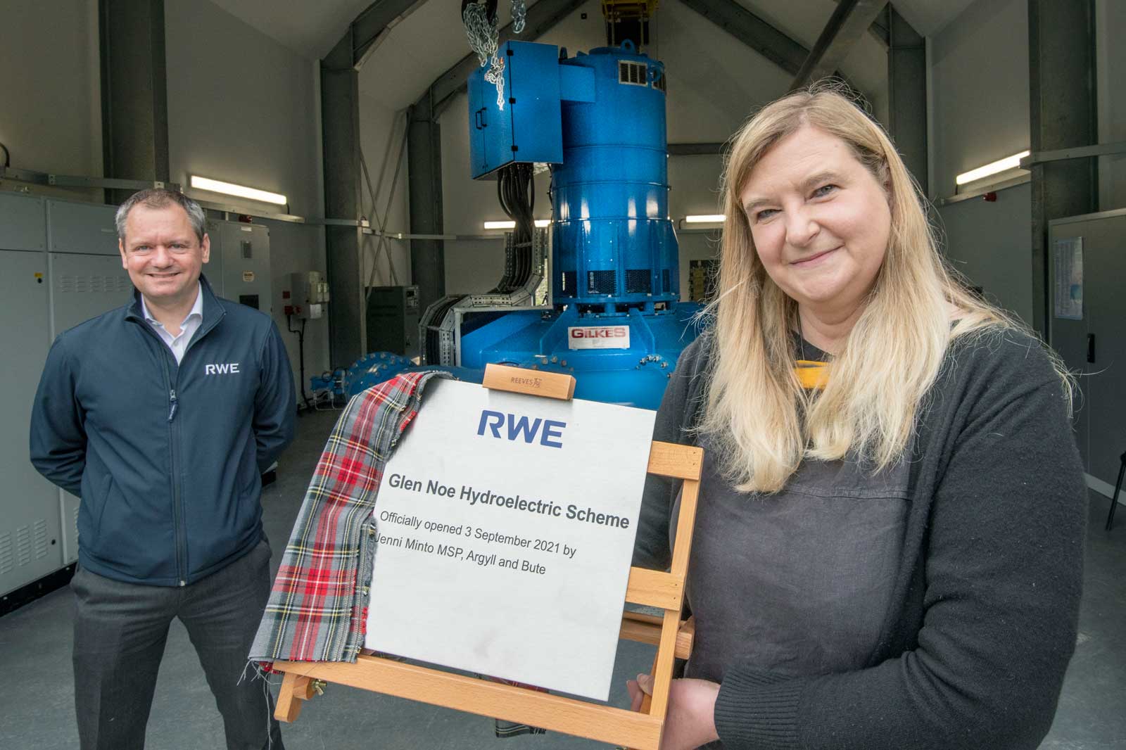 A woman holds a plaque for the Glen Noe Hydroelectric Scheme, with machinery visible in the background.