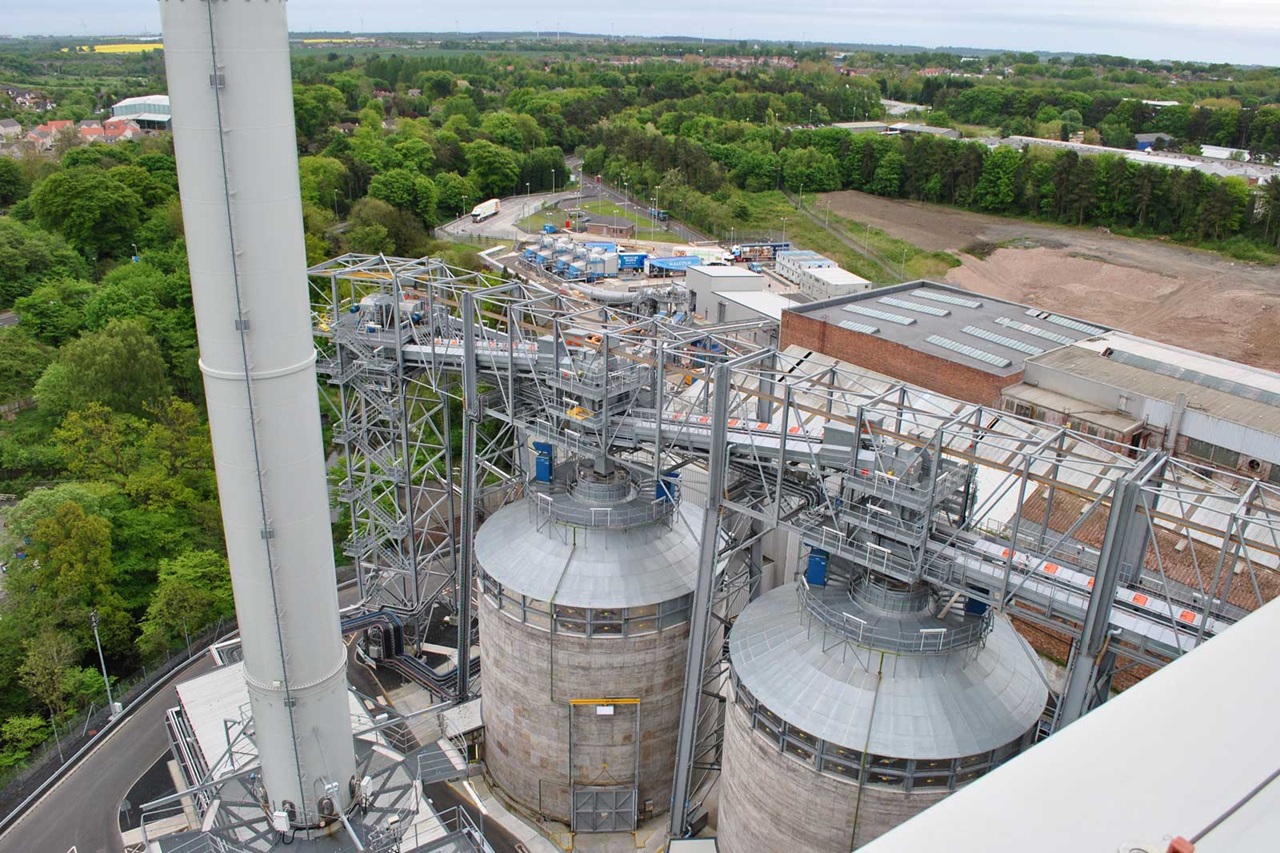 Aerial view of industrial silos and steel structures surrounded by greenery, with nearby construction and buildings visible.