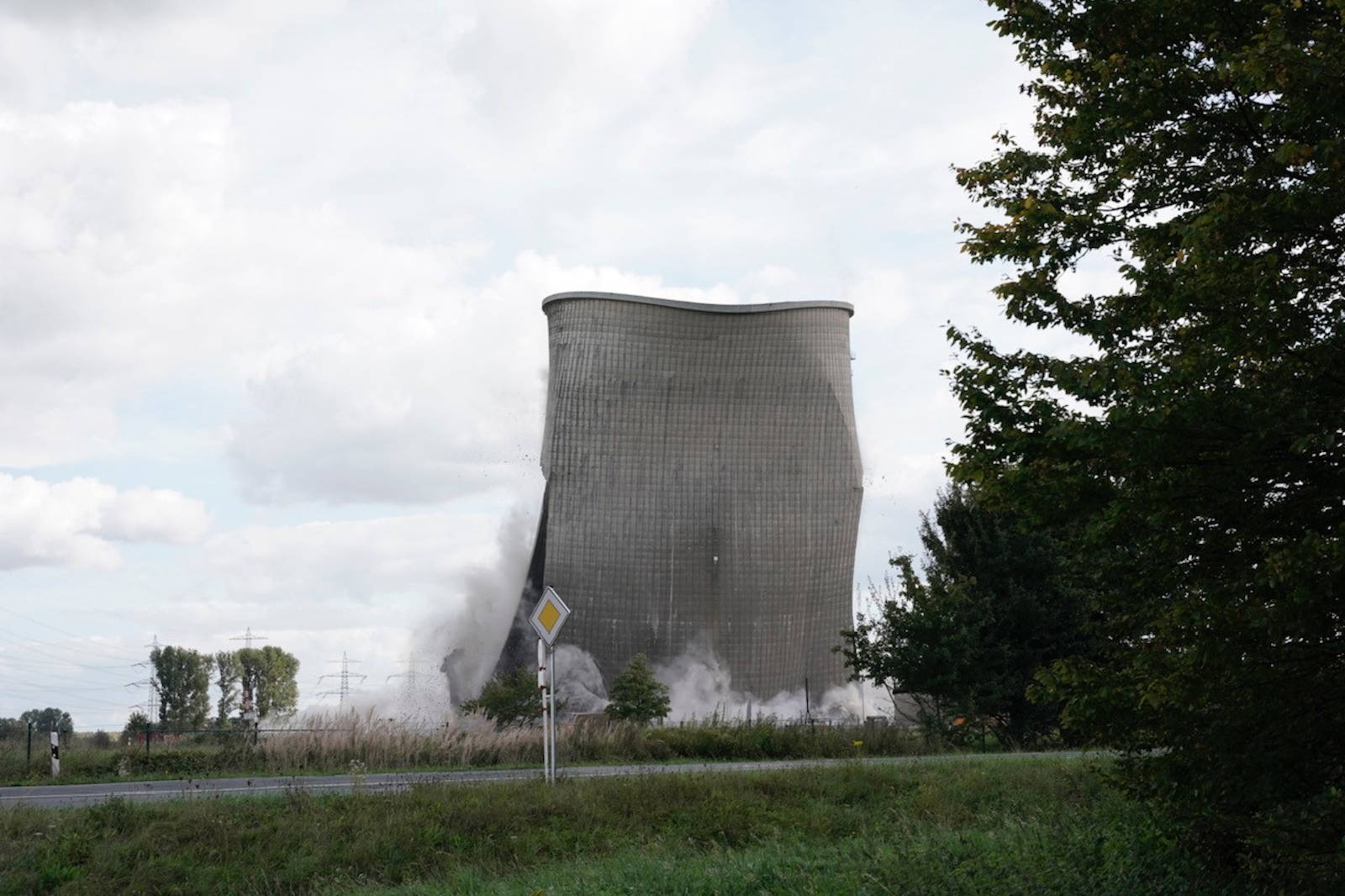 Ein zusammenbrechender Kühlturm mit Staub und Trümmern, umgeben von Grünflächen und Stromleitungen unter einem bewölkten Himmel.