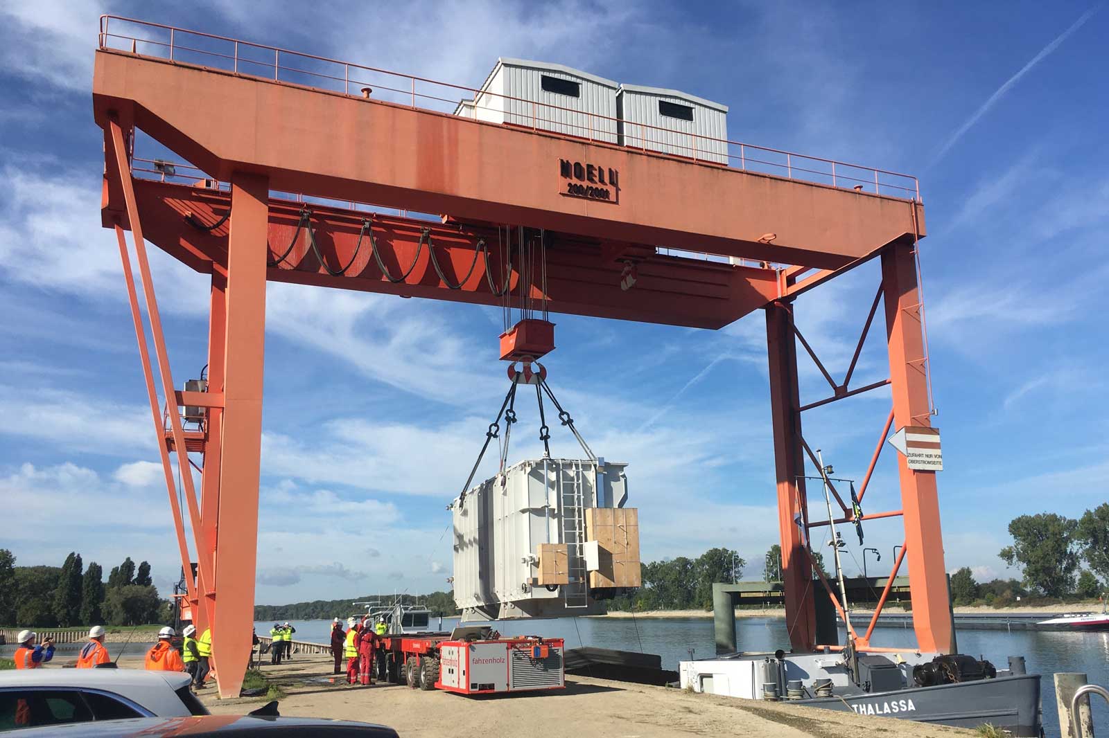 Ein großer oranger Kran hebt einen weißen Container in der Nähe eines Flusses, während Arbeiter zuschauen und ein Boot am Dock ist.