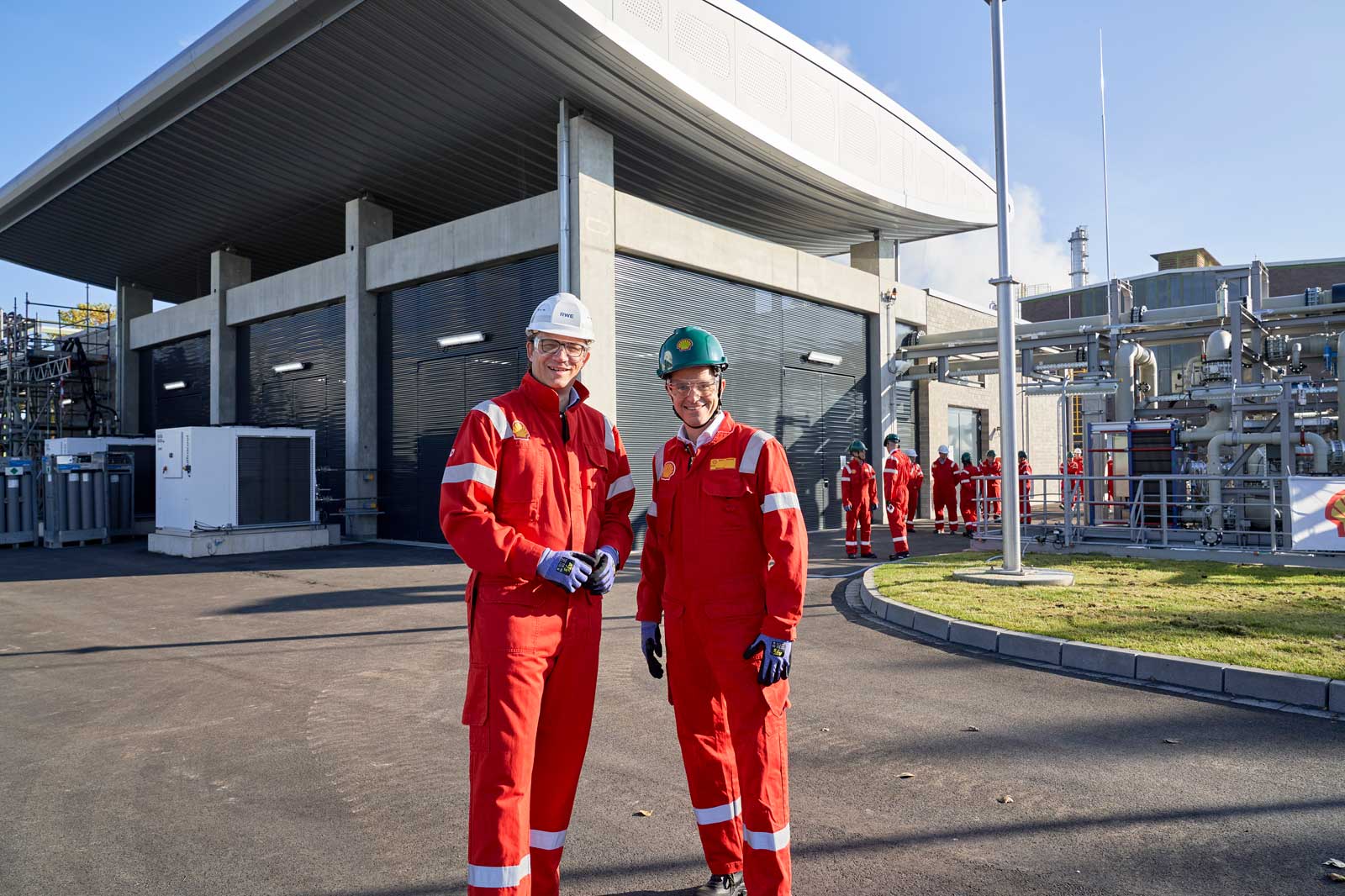 Zwei Arbeiter in roten Uniformen posieren an einer modernen Industrieanlage mit weiteren Arbeitern im Hintergrund.