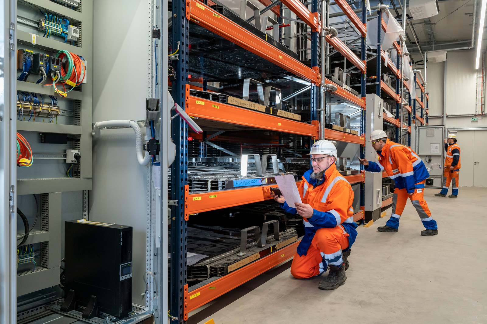 Two workers in orange safety clothing are standing and kneeling in a technical room with storage racks.