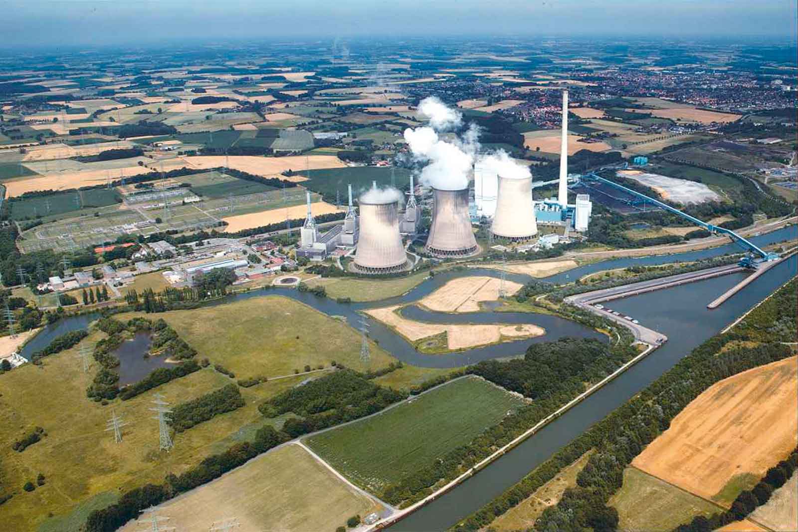 Luftaufnahme eines Kraftwerks mit Kühltürmen, Fluss und landwirtschaftlichen Flächen im Hintergrund.